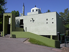 Moscheegebäude mit glatter Betonfassade, hinter einer grünen, niedrigen Mauer