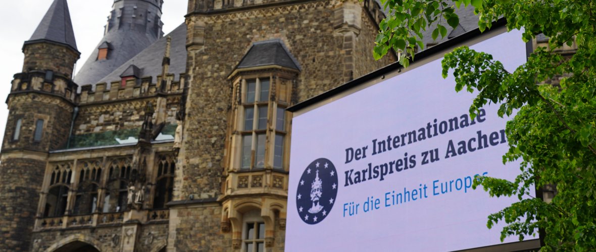A banner with the inscription "The International Charlemagne Prize of Aachen - For the Unity of Europe" hangs in front of the town hall. Green trees protrude into the right-hand edge of the picture.