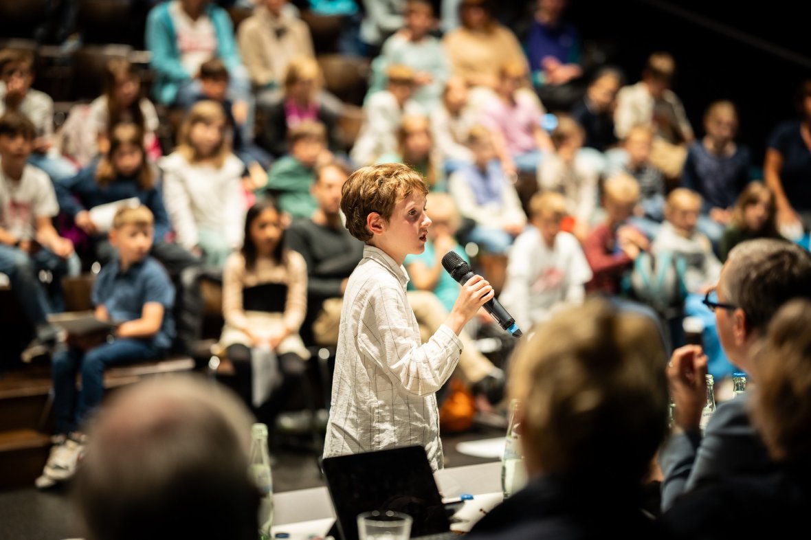 A boy speaks at the event. Other children and the tables with the adults stand around him.