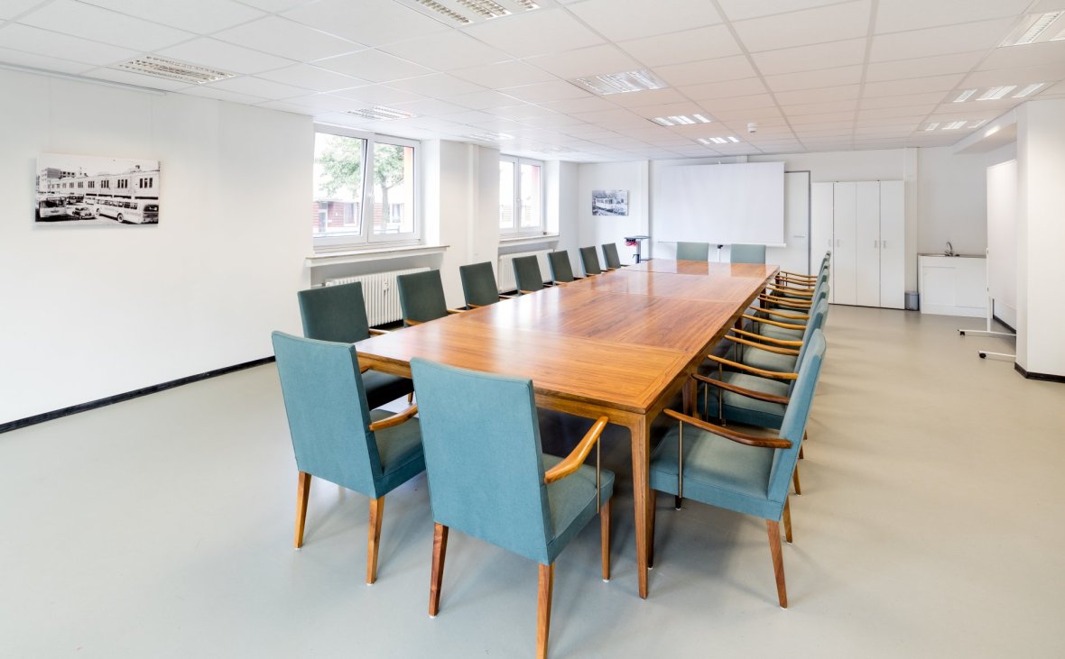 You can see a very bright room with large windows. The shot is very dynamic, so that the conference table can be seen diagonally from left to right in the middle. The table has a warm wood tone. There are 18 chairs around the table. The chairs are upholstered in a beautiful shade of turquoise. In contrast, the wood tone of the table is picked up in the armrests and chair legs. At the front left of the picture is an old black and white photo of the depot on the wall. On the right, at the top in the background, you can still see a flipchart