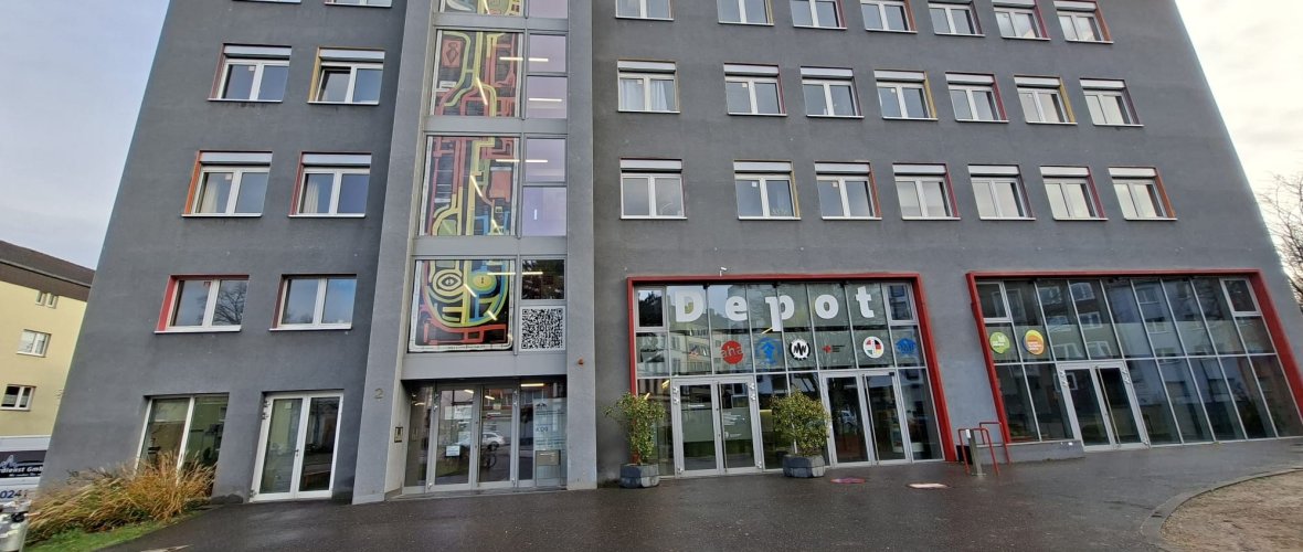 The gray building front of the depot; on the left on the glass front of the stairwell is the artwork with yellow, green, blue and red broad lines on a black background over 4 floors.