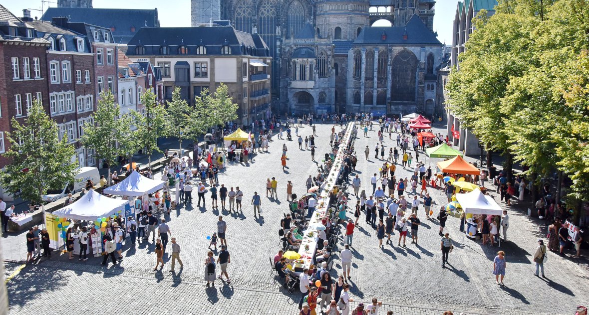Wide view of Aachen's Katschhof with the cathedral in the background from above. The approx. 20-metre "Long Table" stretches out in the foreground of the square. People are sitting along the table, chatting, listening to the music of the stage program and enjoying a meal together. The table is decorated with flowers and parasols and the table is festively laid. Information stands are set up around the table where people can exchange information.