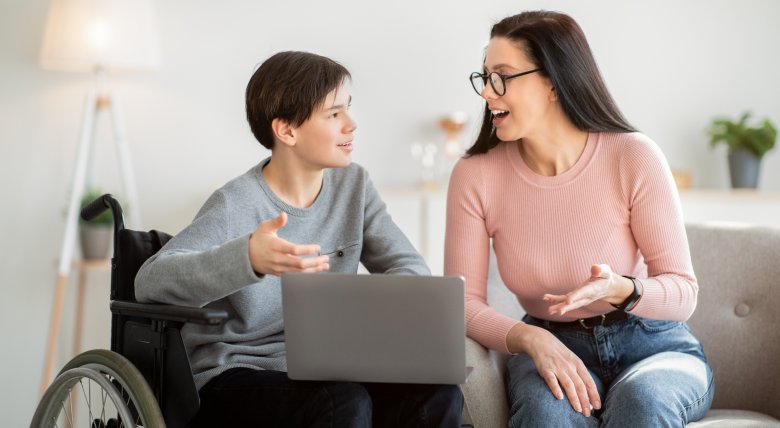 A boy in a wheelchair looks into a laptop with an adult.