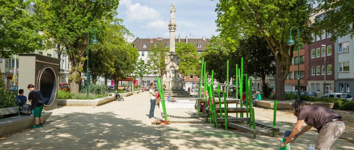 A new playground with climbing structures and plenty of green space. Children are playing; on the right, a woman is helping a toddler onto a floor trampoline.