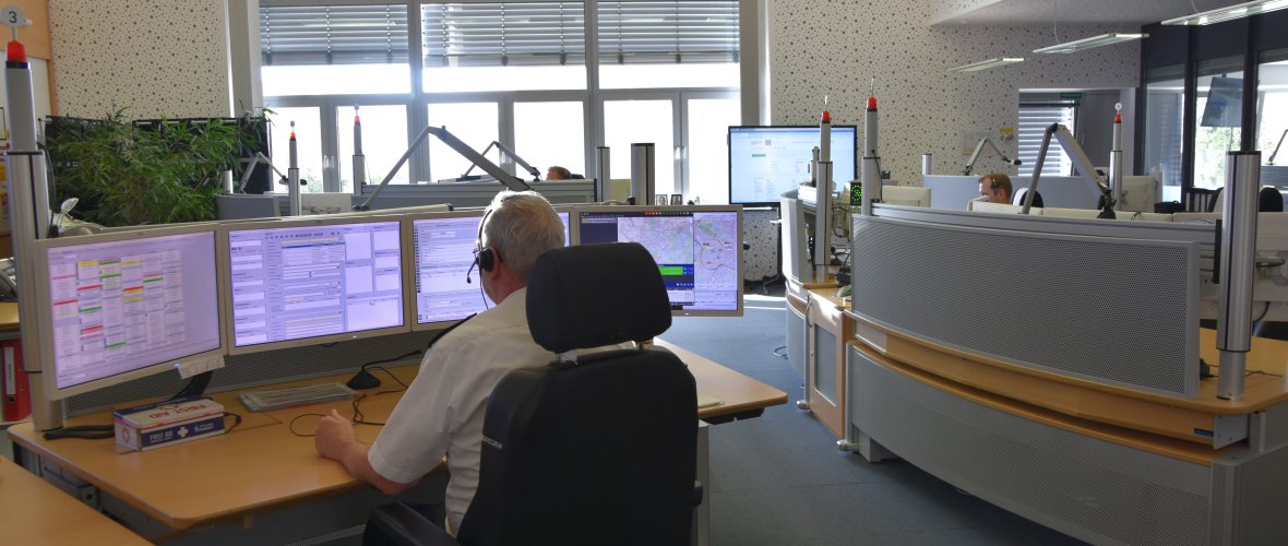 In day-to-day operations, several seats are always occupied in the control center and others are always ready for action. Four employees are currently sitting visibly at their desks and taking emergency calls. One employee is sitting with his back to the camera and has various programs on the screens in front of him. These include a detailed map of Aachen.