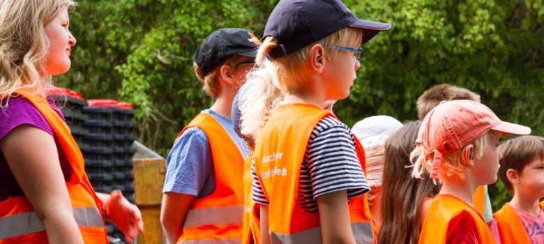 Children in summer clothes and orange vests with "Aachener Stadtbetrieb" printed on them