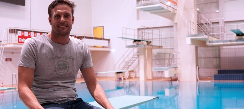 Pool manager Thomas Kelch sits on a diving board at the edge of the pool and smiles at the camera. High diving towers can be seen in the background.