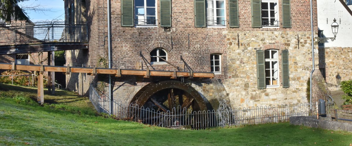 The Welsche Mühle under a blue sky and sunshine. The building is made of old bricks. The green shutters are open.