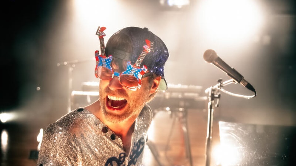 A band member looks open-mouthed at the camera, a microphone next to him. He is wearing sunglasses with electric guitar-shaped lenses and a silver suit.
