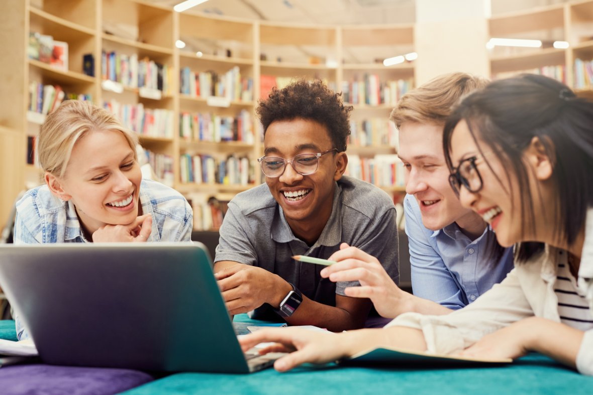 Positively excited students in casual clothes, lying on the floor in the library and laughing while watching a strange video on the laptop.