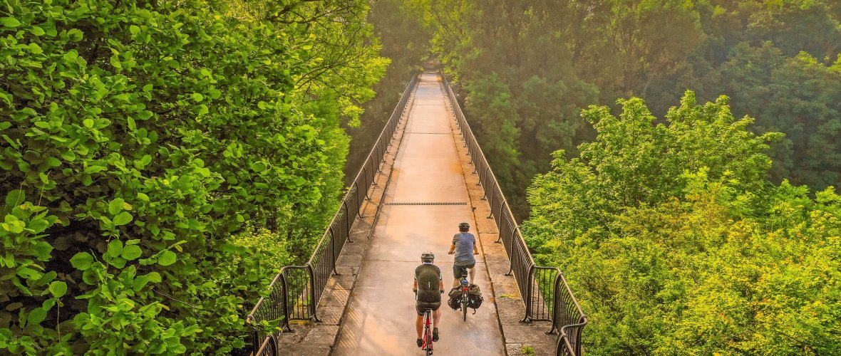 Two people are cycling on a bridge (viaduct) towards the sun. The bridge is surrounded by many tall trees.