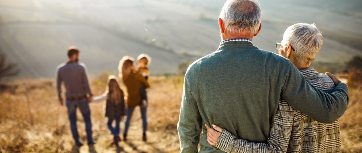 Back view of embraced grandparents looking at their family in a field on a fall day.