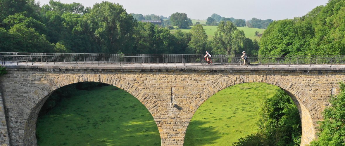 Two cyclists ride over a bridge in the countryside.