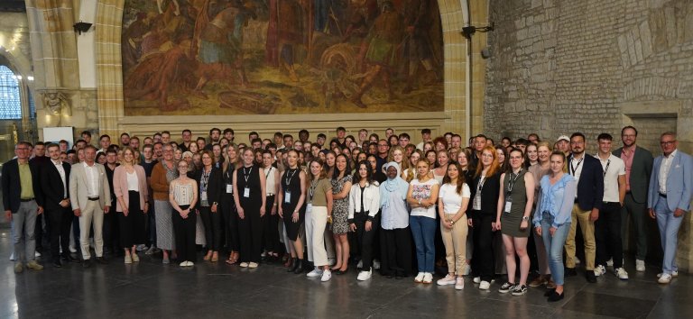 The new trainees of the City of Aachen stand together with Lord Mayor Sibylle Keupen and other responsible persons of the City of Aachen in the Coronation Hall of Aachen City Hall, in the background a large mural painting and the historic masonry of Aachen City Hall.
