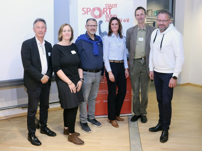 Six people stand in front of a banner with the inscription "Stadt Sport Bund Aachen e.V."