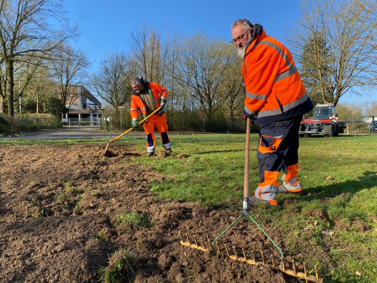 Two men in orange work clothes work the soil with a shovel and rake