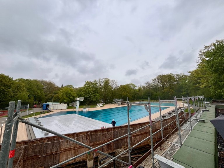 The large pool of the outdoor pool with diving tower can be seen. In the foreground, a construction fence protrudes into the picture.