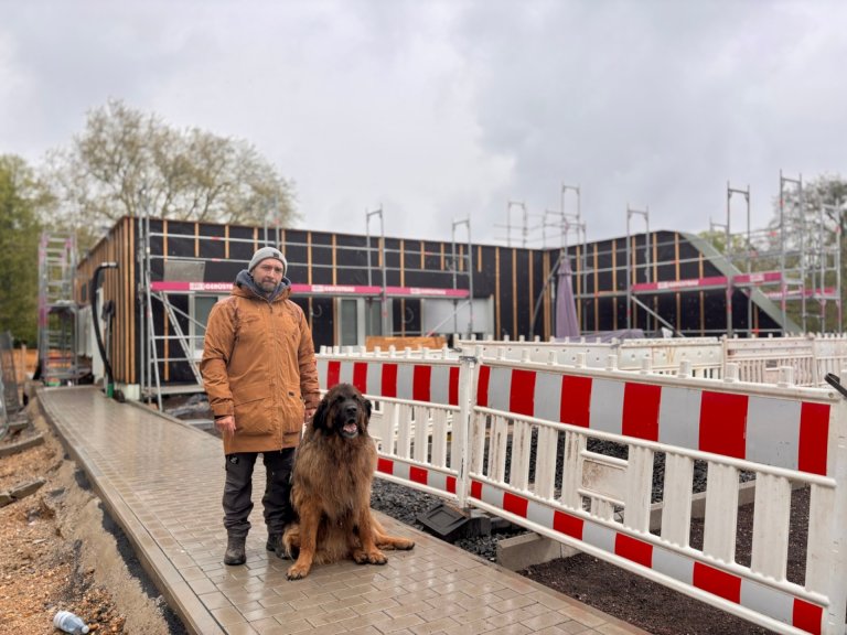 A man stands with a large dog on a newly paved path. A newly built residential building can be seen in the background.