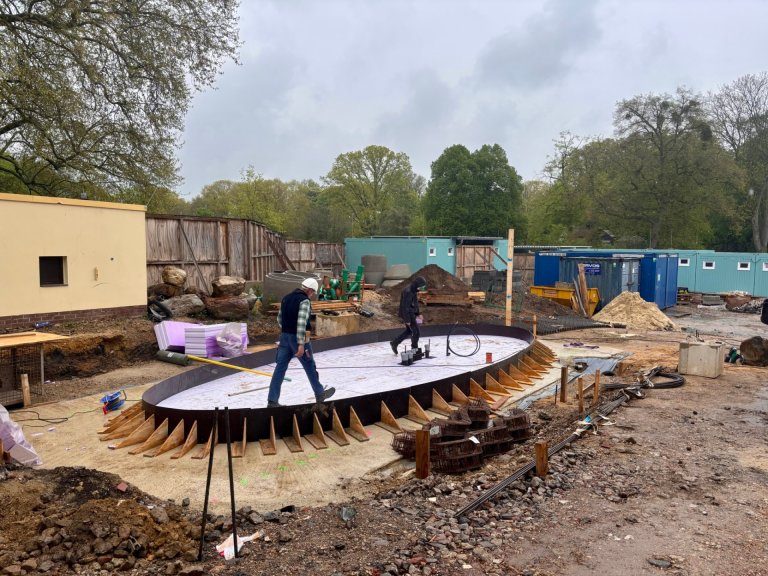 Two construction workers walk across a building site. They are standing on a concrete slab where the new ticket office for the outdoor pool will be built.