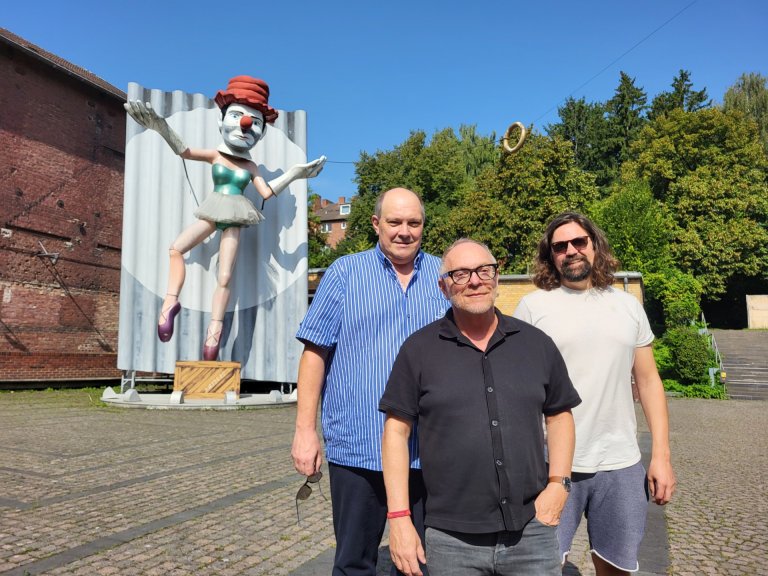 Three men stand in a courtyard. In the background is a larger-than-life sculpture of a dancing clown.