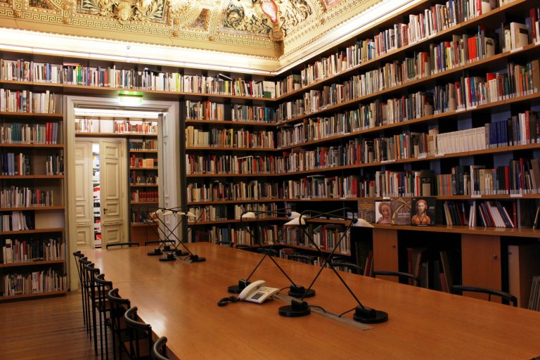 Long wooden tables with reading lamps, chairs in front of them. Shelves with books all around, a white double door. Upper edge of the picture: decorated ceilings.