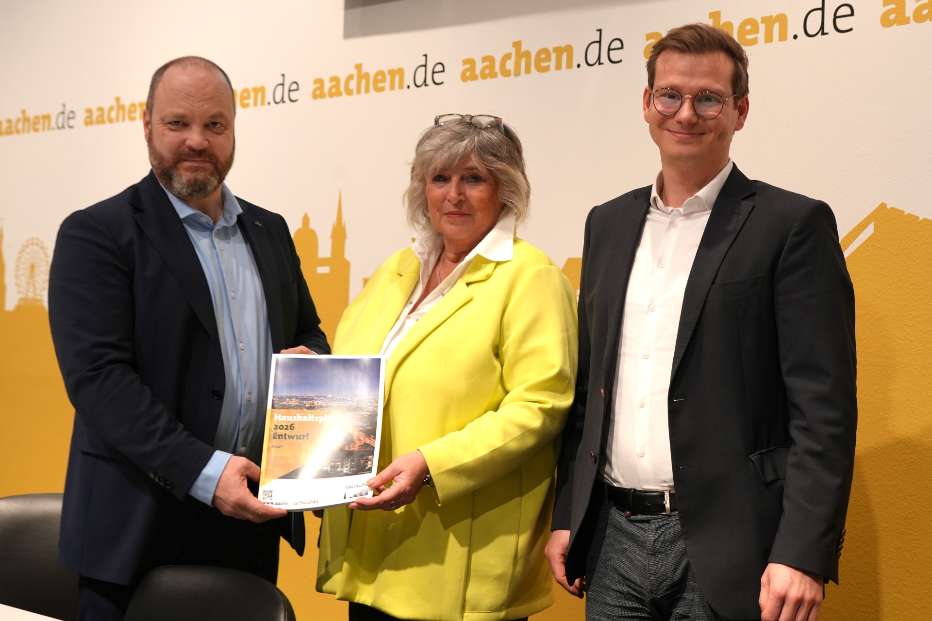 Three people stand in front of a wall with "aachen.de" written on it. The woman in the middle is holding up a brochure with the title "Budget 2026 draft".
