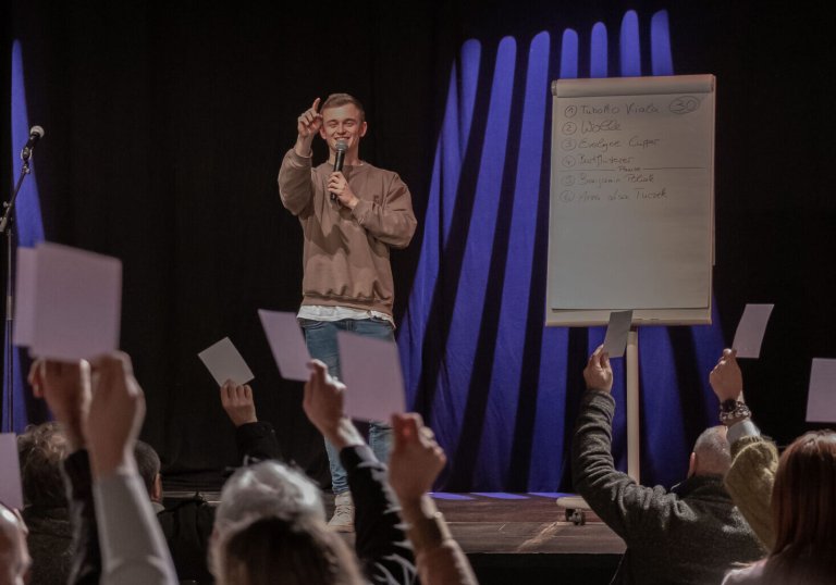 A young man on a stage with a microphone, smiling and pointing towards the audience. The audience holds up white pieces of paper with their hands.