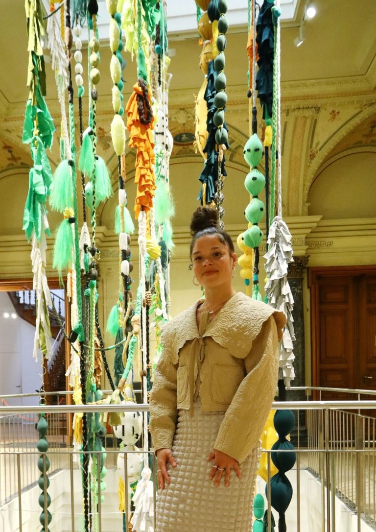 A woman stands in front of a railing and is surrounded by colorful textile strands hanging from the ceiling.