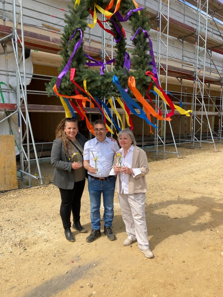 Three people stand under a colorfully decorated wreath and smile