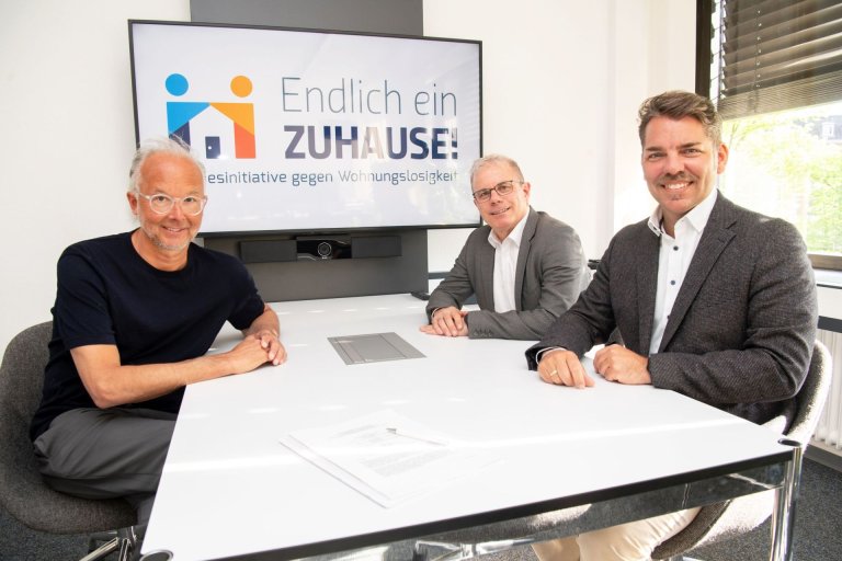 Three men sit at a table in front of the screen with the inscription "Finally a HOME"