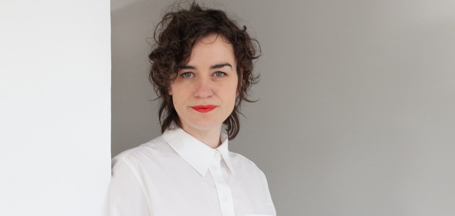 Rebekka Seubert has dark hair and is wearing a white blouse. She is leaning against the wall.