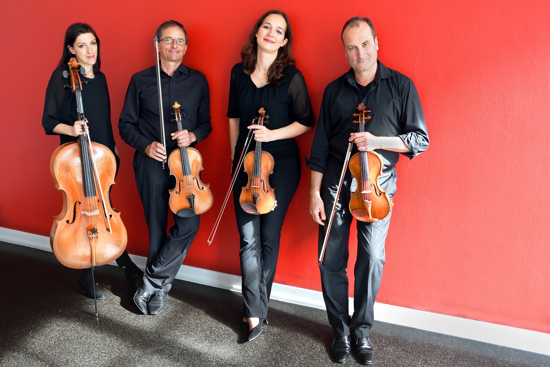 Four people with string instruments in their hands stand in front of a red wall