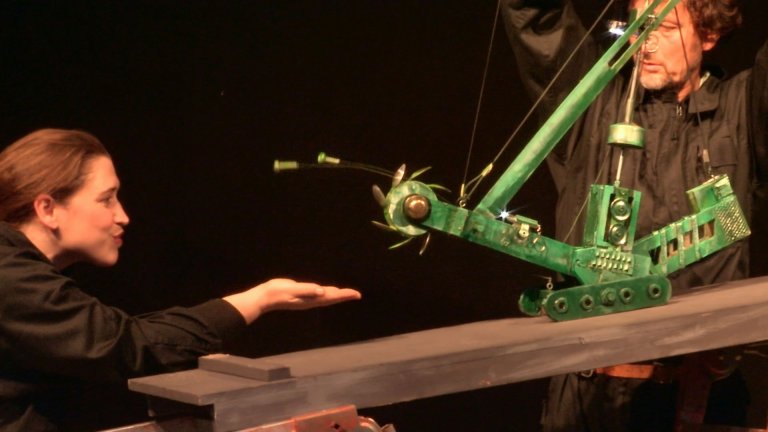 Two people, shown from the side, are standing next to a green metal frame. The man on the right is holding the frame, the woman on the left is reaching out her hand towards the frame.