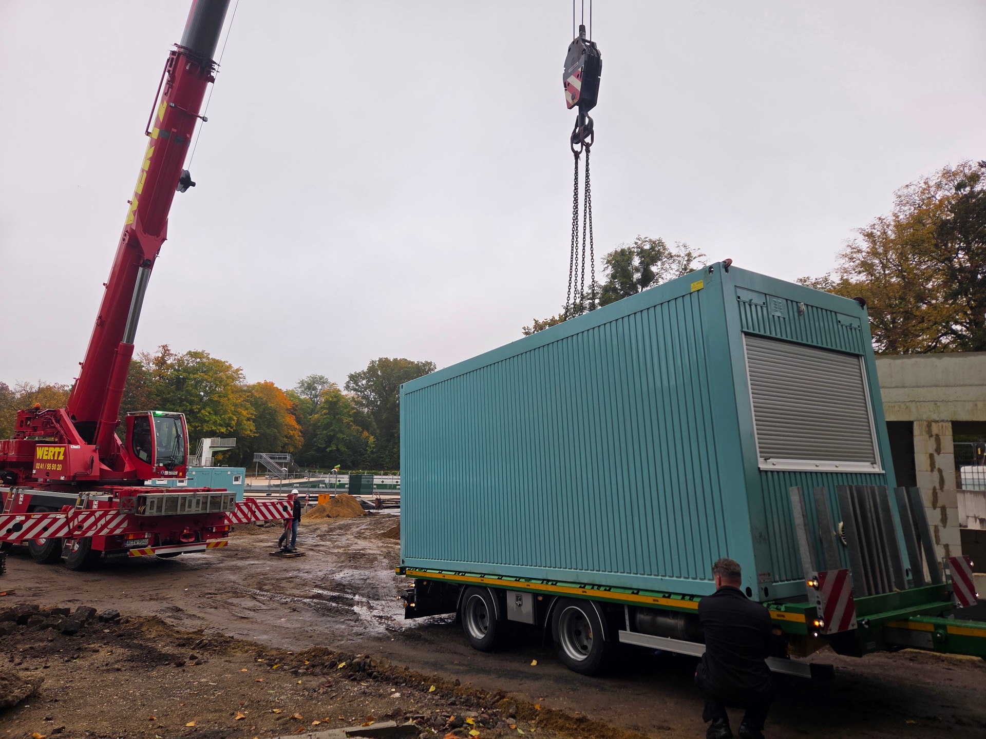 A large red crane lifts a colored container onto a truck. The vehicles are parked on the construction site at the Hangeweiher outdoor pool.