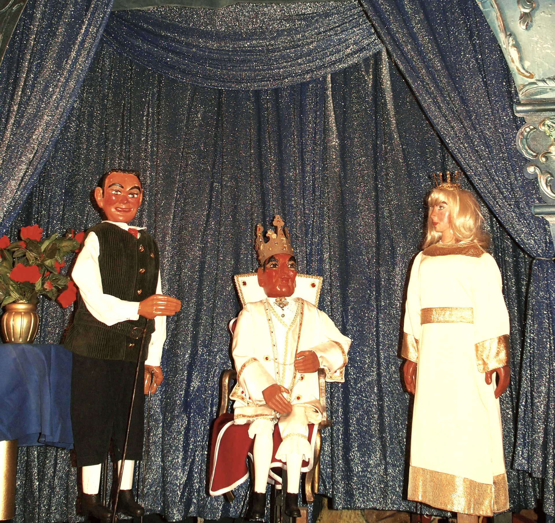 Three puppet figures in front of a glittering curtain: a male figure standing on the left, a king with a crown sitting on a throne in the middle and a woman with a small crown and a white dress standing on the right.