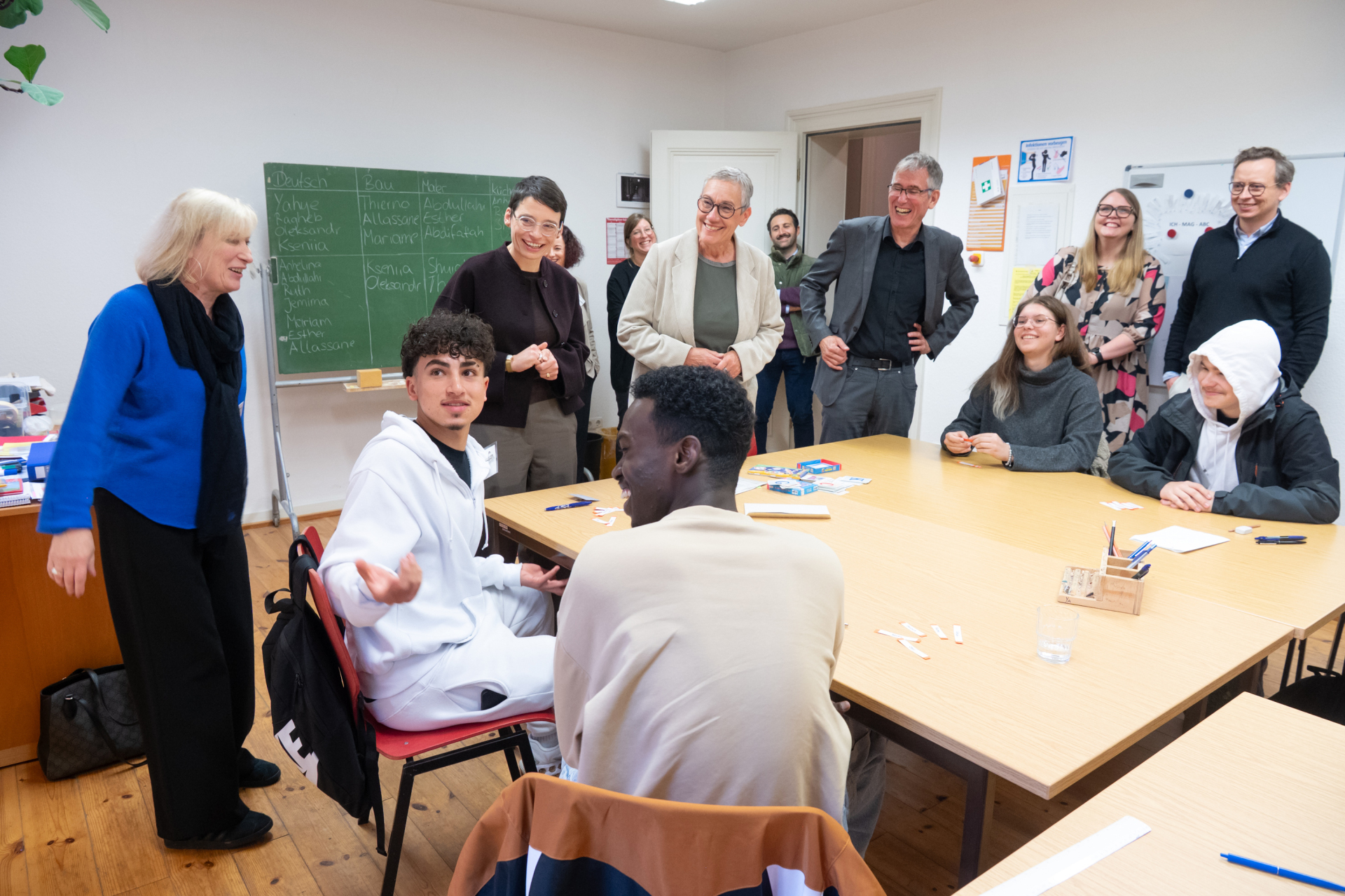 A classroom with four pupils sitting at a table. People laughing and a blackboard with writing on it in the background.