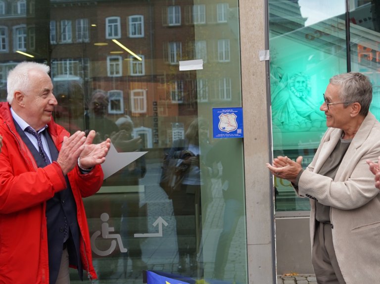 A man (left) and a woman (right) clap. A blue plaque hangs on a glass front between them