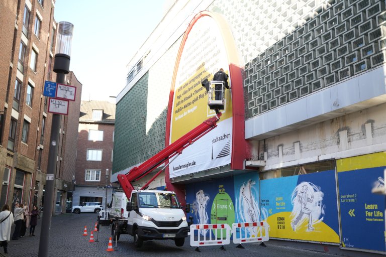 A large banner is attached to the façade of the former department store by workmen using a lifting platform.