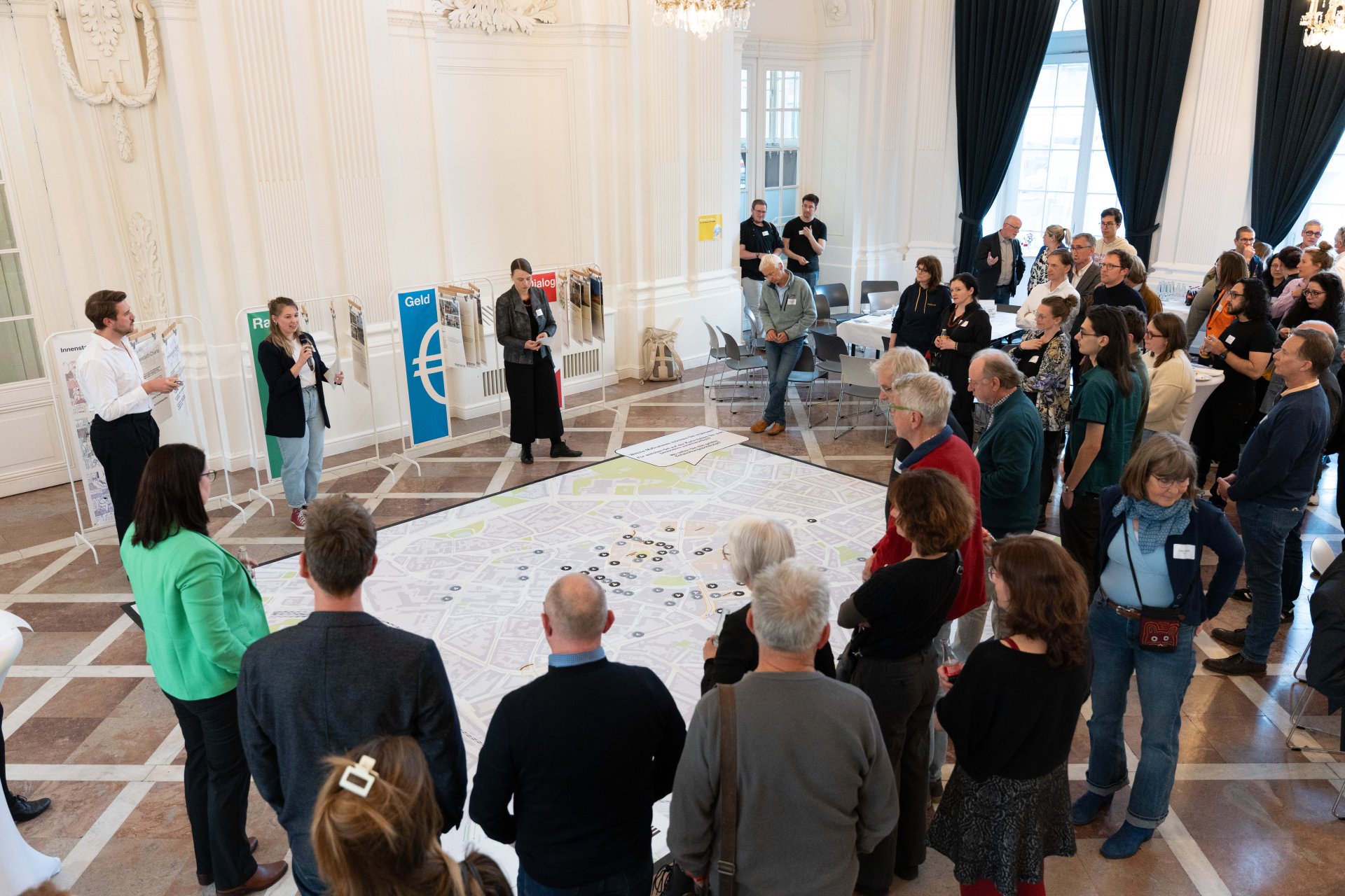 A group of people are standing in a room. There is a large map of the city on the floor.