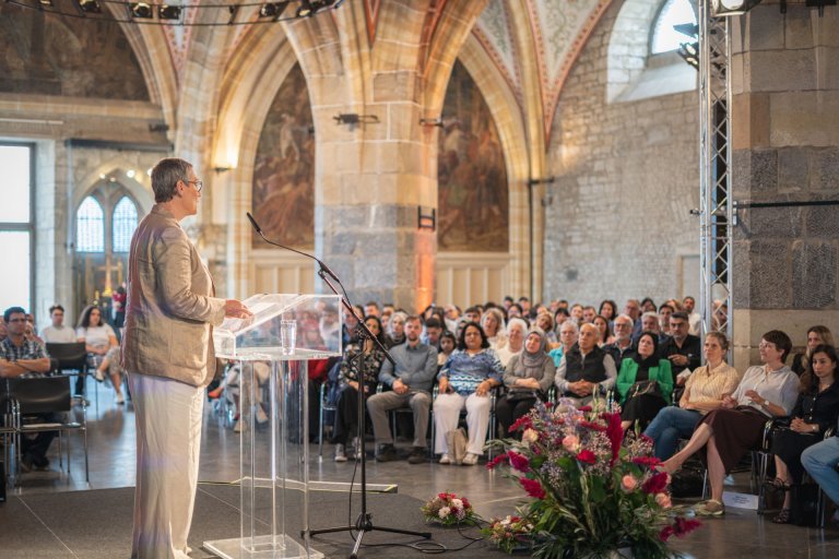 A woman stands at a lectern with her back to the camera. She is speaking to many people sitting in a large, historic hall.