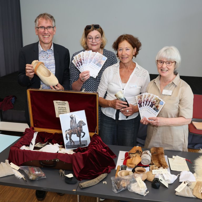 Four people are standing in front of an open suitcase lying on a table. Around and inside the suitcase are various objects relating to Charlemagne.