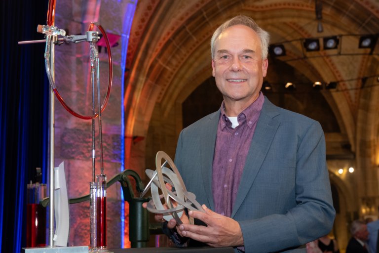 A man smilingly holds a metal sculpture of intertwined ellipses in his hand.