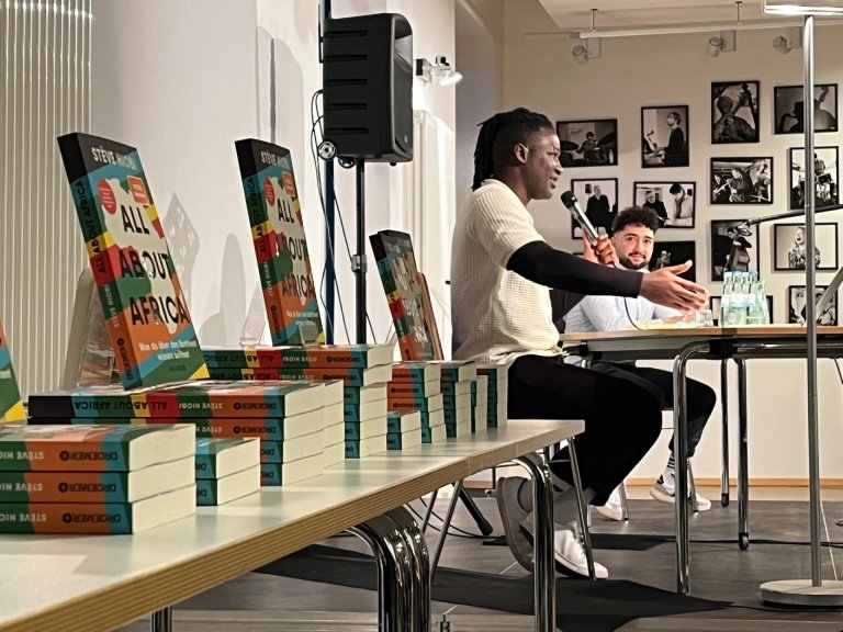 A man sits on a stage and speaks to an audience. A table with books in the left section of the picture.