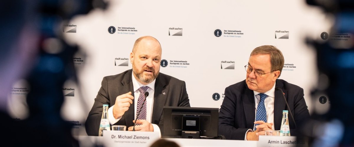 Two men sit on a podium under a picture of another man.