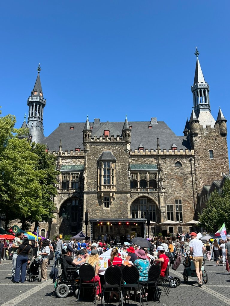 In a square stands a table dozens of meters long, covered with white tablecloths. People are standing and sitting all around it. In the background is a stage set up in front of Aachen’s historic city hall.
