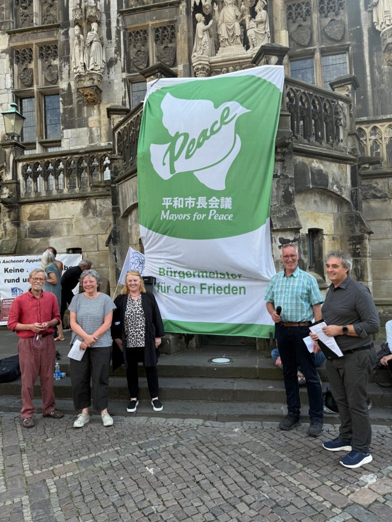 A group of people is standing in front of a green-and-white flag on the steps of City Hall.