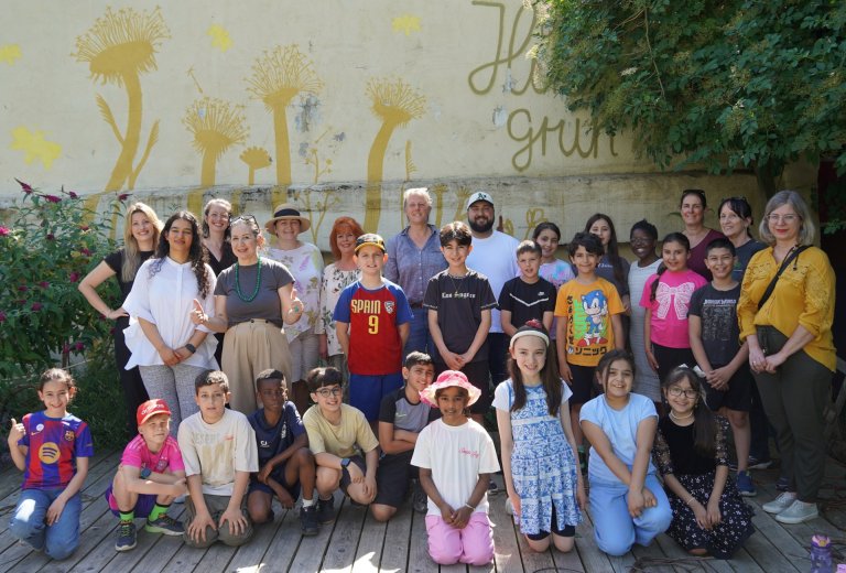 A group of people, with two rows of children in the front, are standing together in front of a wall painted with flowers.
