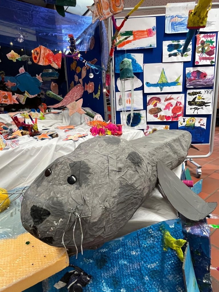 A large seal sculpture in the foreground, with colorful pictures and handmade fish hanging from the ceiling in the background.