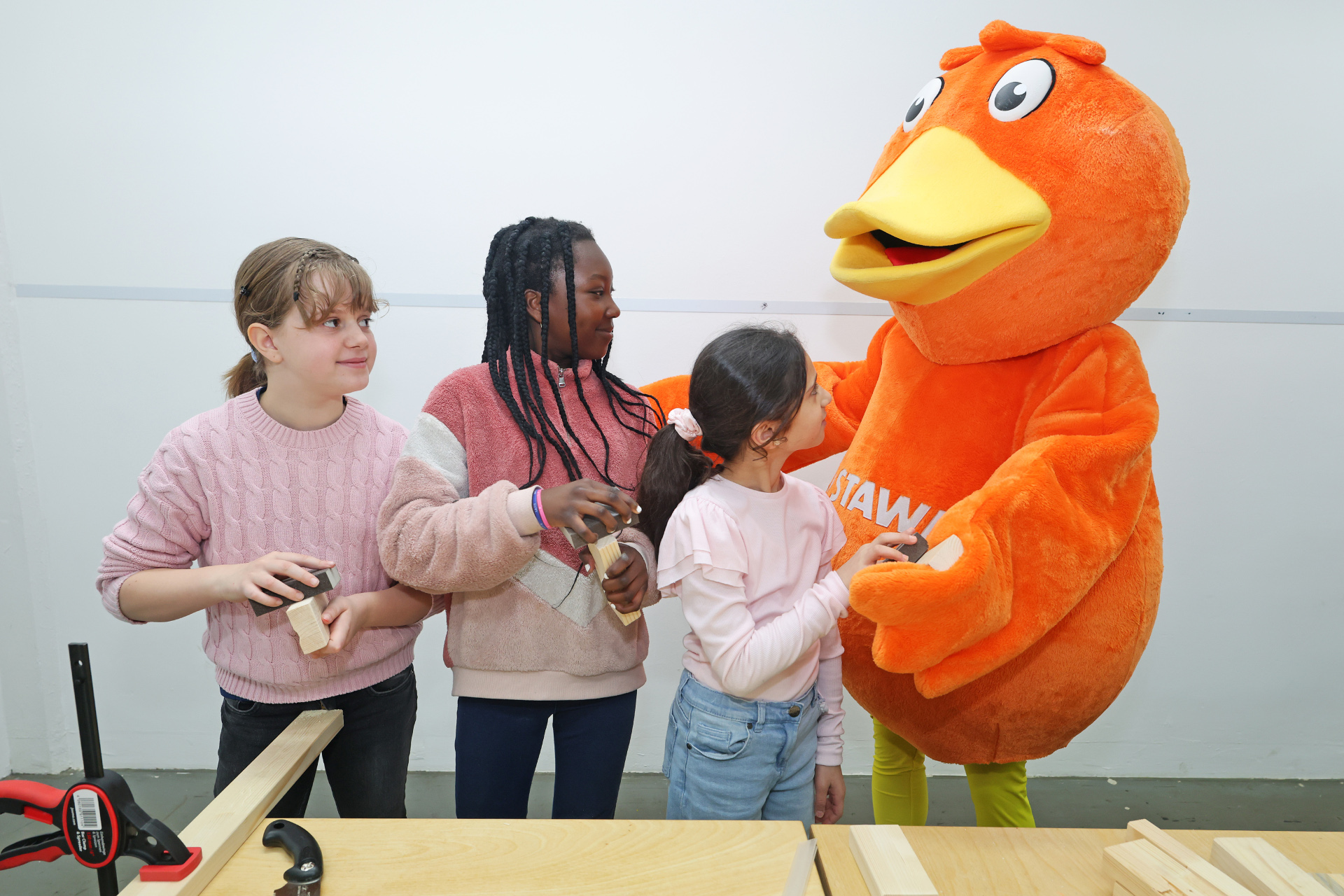 Three girls are standing at a workbench sanding pieces of wood, with STAWAG's mascot, an orange duck, standing next to them.