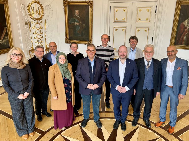 A group of people stand in the middle of a room with white, gold-decorated walls and look into the camera.